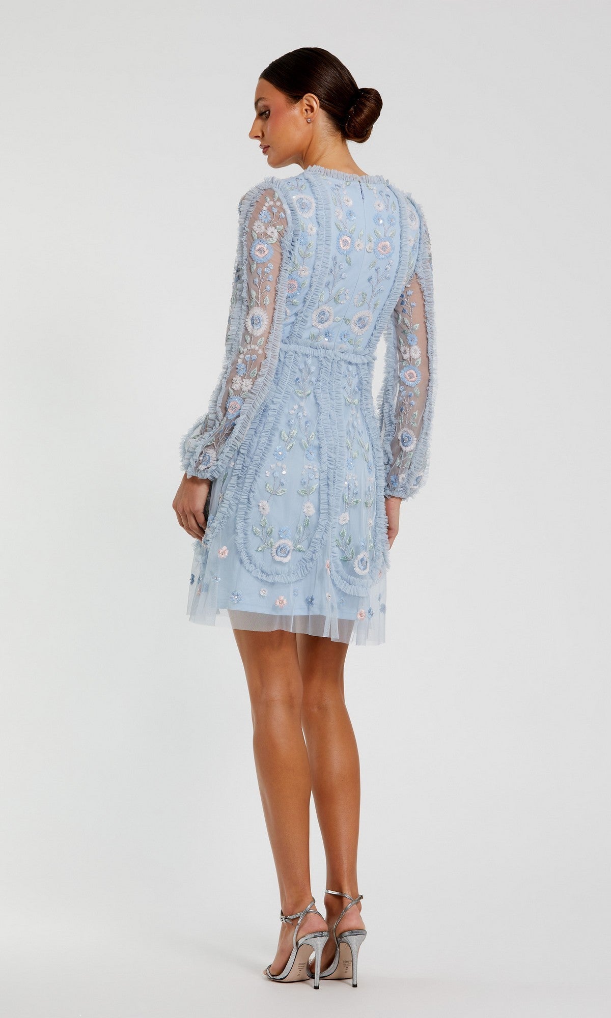 A woman faces away, showcasing the Mac Duggal 9365 Short Wedding Guest Dress in light blue with long sleeves and floral embroidery, paired with silver high-heeled sandals against a plain background.